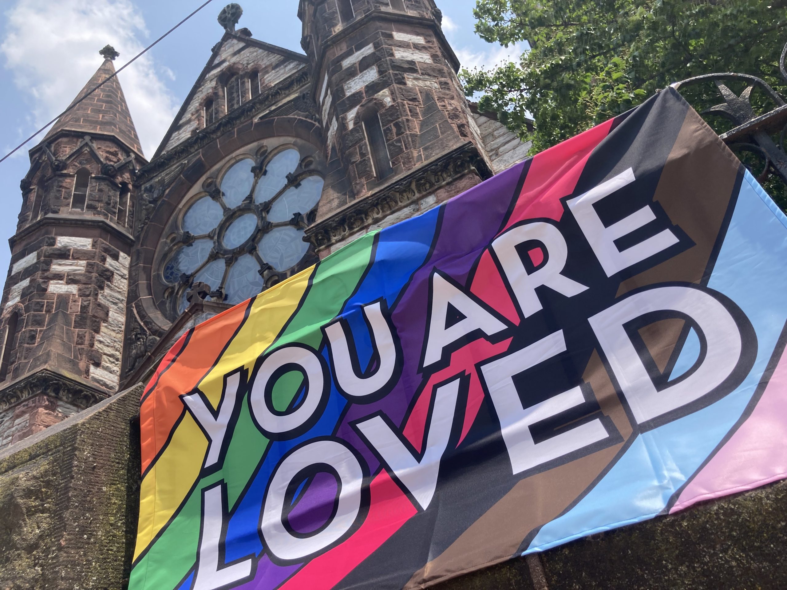 exterior of a stone church with a rainbow banner that says "You Are Loved"